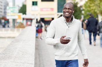 Black young man with a smartphone in his hand in urban background. Young african guy with shaved head wearing casual clothes and white headphones.