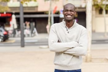 Black young man with arms crossed smiling in urban background. Young african guy with shaved head wearing casual clothes.