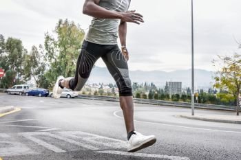 Black man running outdoors in urban road listening to music with white headphones. Young male exercising with city scape at the background.