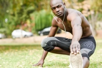 Fit shirtless young black man doing stretching before running in urban park. Young male exercising with naked torso listening to music with headphones.