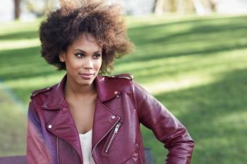 Young black female with afro hairstyle sitting in a bench in an urban park. Mixed woman wearing red leather jacket and white dress with city background.