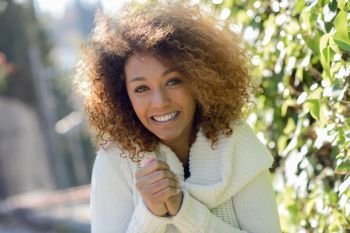 Beautiful young African American woman with afro hairstyle and green eyes wearing white winter dress with cold face