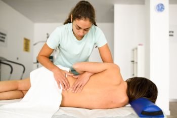 Professional female physiotherapist giving lumbar shoulder massage to brunette woman in hospital. Medical check at the shoulder in a physiotherapy center.