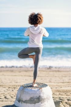 Rear view of young black woman doing yoga in the beach. Female wearing white sport clothes in tree asana with defocused sea background. Afro hairsytle.