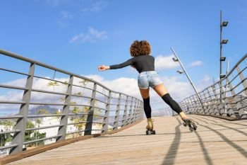 Rear view of black woman, afro hairstyle, on roller skates riding outdoors on urban bridge. Young girl rollerblading on sunny day.