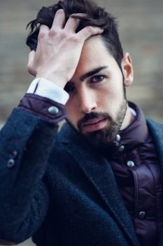 Young bearded man, model of fashion, in urban background wearing british elegant suit. Guy with beard and modern hairstyle touching his hair