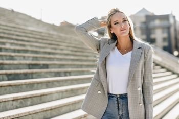 Beautiful young caucasian woman in urban background. Blond girl wearing casual clothes in the street. Female with elegant jacket and blue jeans standing on stairs.