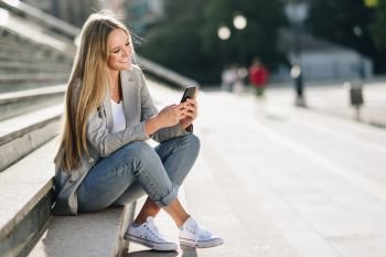 Beautiful young caucasian woman looking at her smartphone and smiling in urban background. Blond girl wearing casual clothes sitting on stairs.