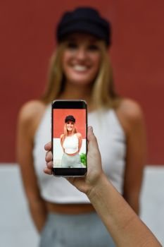 Woman hand taking photograph with a smartphone to her blonde friend outdoors. Model wearing white t-shirt and cap.