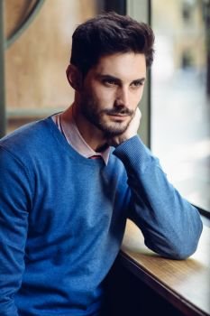 Thoughtful man with blue sweater with lost look near a window in a modern pub. Bearded guy with modern hairstyle. Life style concept.