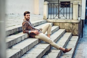 Young smiling man, model of fashion, wearing modern shirt. Guy with beard and modern hairstyle sitting in urban steps.