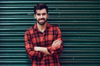 Young smiling man, model of fashion, wearing a plaid shirt with a green blind behind him. Guy with beard and modern hairstyle in urban background.