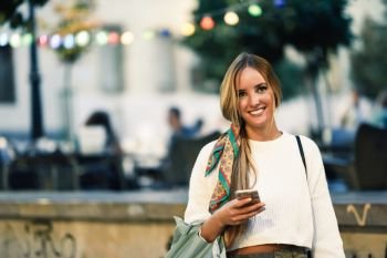 Blonde woman with smartphone in the street. Defocused city lights at the background. Pretty girl with pigtail hairstyle at night.
