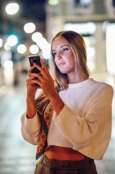 Blonde woman taking photograph with smartphone at night in the street. Defocused city lights at the background. Pretty girl with pigtail hairstyle at night.