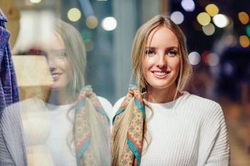 Blonde girl wearing white sweater smiling in the street with defocused city lights at the background. Pretty woman with pigtail hairstyle at night reflected in a shop window.