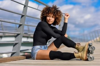Black woman, afro hairstyle, on roller skates sitting outdoors on urban bridge. Young female with beautiful clouds at the background.