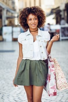 Young black woman, afro hairstyle, with shopping bags in the street. Girl wearing casual clothes in urban background. Female with skirt, denim vest and high heels.