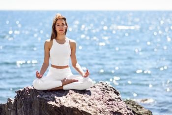 Young woman doing yoga in the beach. Female wearing white sport clothes in lotus figure with defocused sea background.