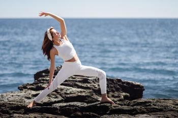 Young woman doing yoga in the beach. Female wearing white sport clothes in warrior at sun figure with defocused sea background.