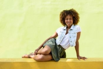Young black woman, afro hairstyle, sitting on a wall smiling. Girl, model of fashion, wearing casual clothes in urban background. Female with skirt, denim vest and high heels.