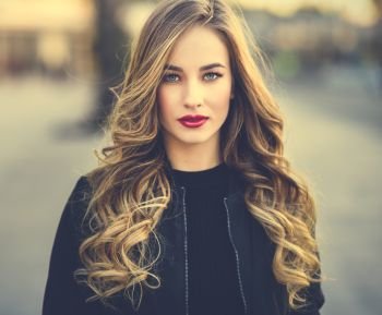 Close-up portrait of young blonde girl with beautiful blue eyes wearing black jacket outdoors. Pretty russian female with long wavy hair hairstyle. Woman in urban background. 
