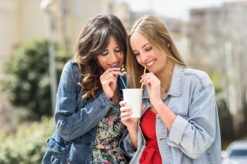 Two young women drinking the same take away glass together with two straws outdoors.