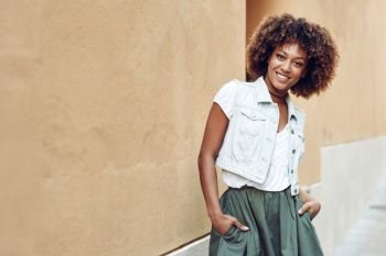 Young black woman, afro hairstyle, smiling near a wall in the street. Girl wearing casual clothes in urban background.