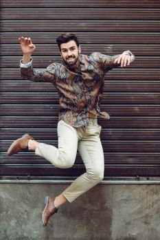 Young bearded man jumping in urban background with open arms wearing casual clothes. Guy with beard and modern hairstyle in the street.