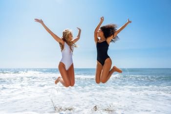 Two funny girls in swimsuit jumping on a tropical beach. Two funny girls with beautiful bodies in swimwear jumping on a tropical beach. Funny caucasian and arabic females wearing black and white swimsuits.