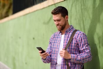 Young man looking at his smart phone in urban background. Lifest. Young man looking at his smart phone and smiling in urban background. Guy wearing casual clothes. Lifestyle concept.