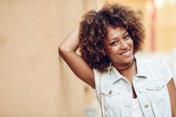 Young black woman, afro hairstyle, smiling in urban background. Young black woman, afro hairstyle, smiling near a wall in the street. Girl wearing casual clothes in urban background.