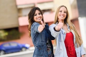 Two young women with thumbs up outdoors. Two funny female friends with thumbs up and looking at the camera in urban background.