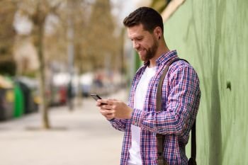 Smiling young man looking his smart phone in urban background. L. Smiling young man looking his smart phone in urban background. Guy wearing casual clothes. Lifestyle concept.