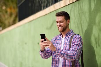 Young man looking at his smart phone in urban background. Lifest. Young man looking at his smart phone and smiling in urban background. Guy wearing casual clothes. Lifestyle concept.