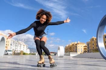 Black woman on roller skates riding outdoors on urban street. Young fit black woman on roller skates riding outdoors on urban street with open arms. Smiling girl with afro hairstyle rollerblading on sunny day