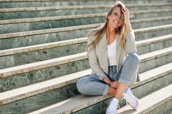 Beautiful young blonde woman smiling on urban steps.. Beautiful young caucasian woman smiling in urban background. Blond girl wearing casual clothes in the street. Female with elegant jacket and blue jeans sitting on stairs.