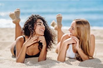 Two young women with beautiful bodies in swimsuit on a tropical . Two young women with beautiful bodies in swimwear on a tropical beach. Funny caucasian and arabic females wearing black and white swimsuits lying on the sand on the beach