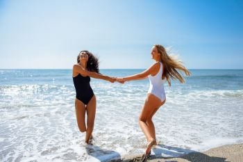 Two women in swimsuit having fun on the beach. Two young women with beautiful bodies in swimwear having fun with their hands caught on the beach. Funny caucasian and arabic females wearing black and white swimsuits