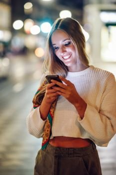 Woman with defocused urban city lights looking at her smartphone. Blonde woman looking at her smartphone in the street. Defocused city lights at the background. Pretty girl with pigtail hairstyle at night.