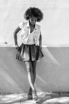 Young black woman, afro hairstyle, standing in urban background. Young black woman, afro hairstyle, standing in the street. Girl wearing casual clothes smiling in urban background. Female with skirt, denim vest and high heels.