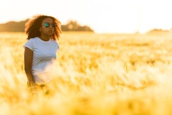 A beautiful mixed race African American female teenager young woman standing in a wheat field at sunset in golden sunshine wearing sunglasses and white t-shirt