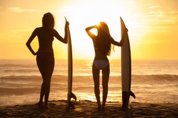 Rear view of two beautiful sexy young woman surfer girls in bikinis with white surfboards on a beach at sunset or sunrise