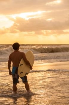 Rear view of young man male surfer with white surfboard looking at surf on a beach at sunset or sunrise