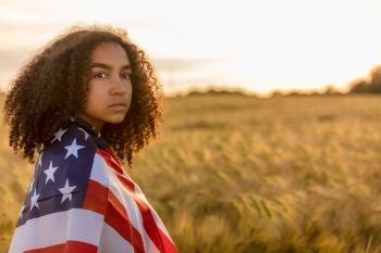 Sad depressed mixed race African American girl teenager female young woman in a field of wheat or barley crops wrapped in USA stars and stripes flag in golden sunset evening sunshine