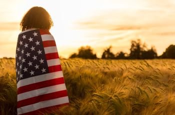 Mixed race African American girl teenager female young woman in a field of wheat or barley crops wrapped in USA stars and stripes flag in golden sunset evening sunshine