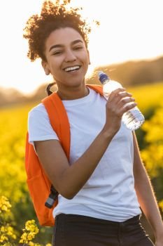 Outdoor portrait in golden evening sunshine of beautiful happy mixed race African American girl teenager female young woman smiling laughing with perfect teeth hiking with orange rucksack and drinking water bottle in field of yellow flowers