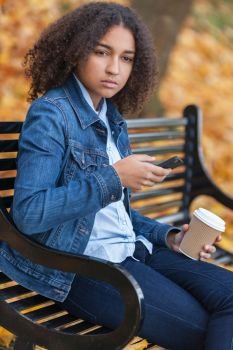 Beautiful mixed race African American girl teenager female young woman using cell phone and drinking takeaway coffee outside sitting on a park bench in autumn or fall looking sad depressed or thoughtful
