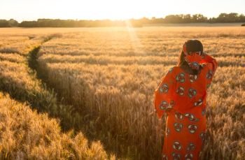 African woman in traditional clothes standing, looking to the sun, hand to eyes, in field of barley or wheat crops at sunset or sunrise