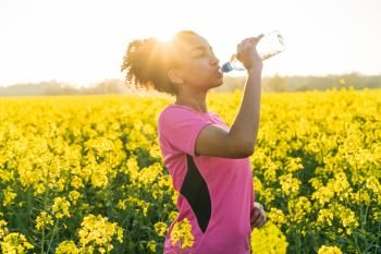 Outdoor portrait of beautiful happy mixed race African American girl teenager female young woman athlete runner drinking water from a bottle in a field of yellow flowers at sunset in golden evening sunshine. Mixed Race African American Girl Teenager Runner Drinking Water 
