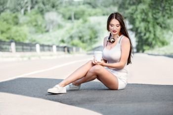 Collage of a beautiful young woman with music headphones around her neck, drinking coffee from a takeaway coffee cup and sitting on the road near a separating strip.